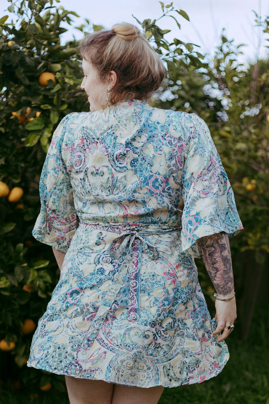 Person wearing a floral dress standing in an outdoor setting with greenery and fruit trees.