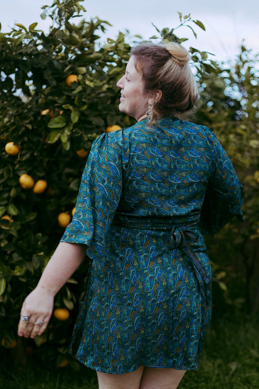 Woman in a blue patterned dress standing in an orchard with fruit trees.