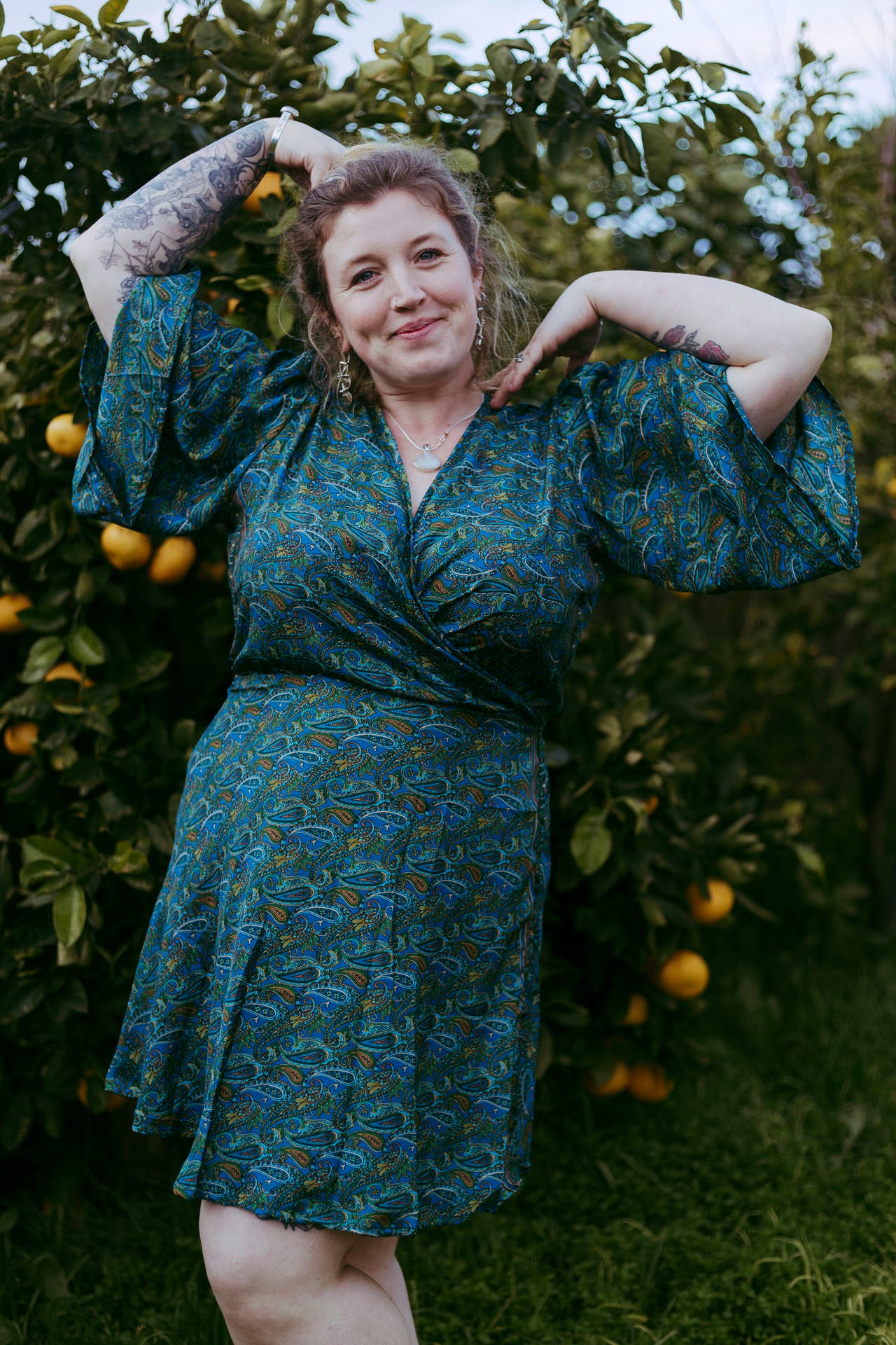 Woman in a blue patterned dress standing outdoors with greenery and fruit in the background