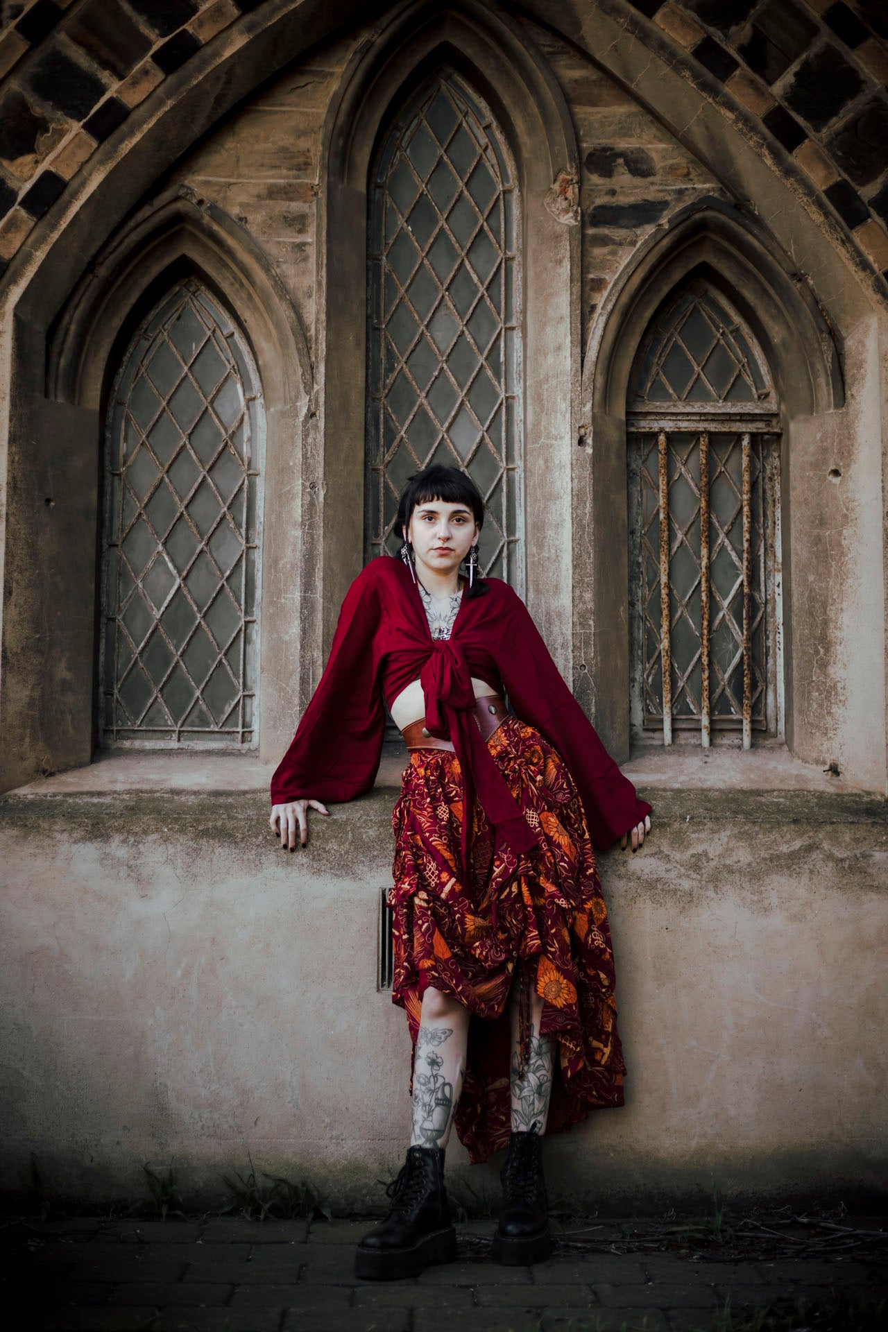 Person in a burgundy coat and floral skirt standing in front of gothic-style windows.