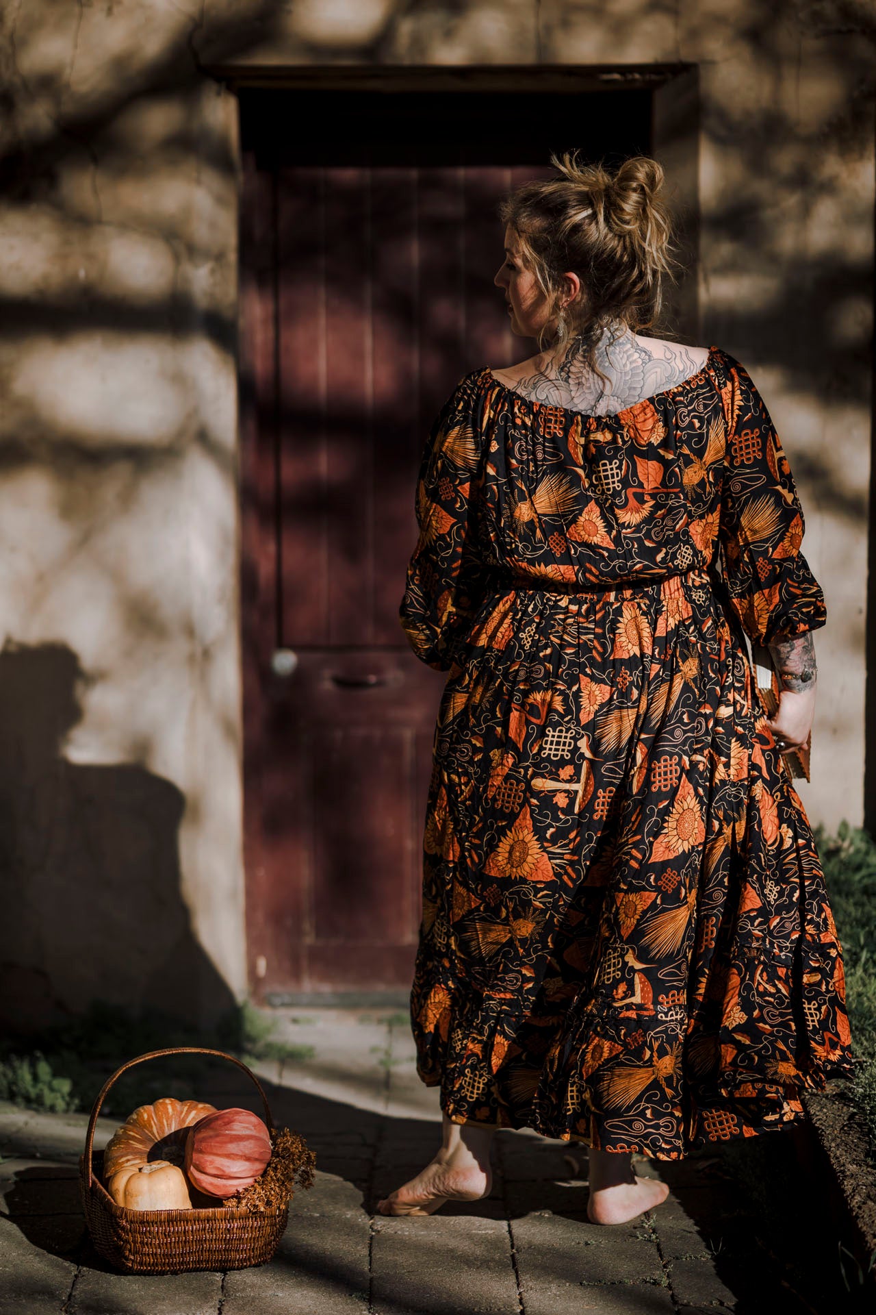Woman in a patterned dress standing in front of a stone wall with a basket of bread and pumpkins.