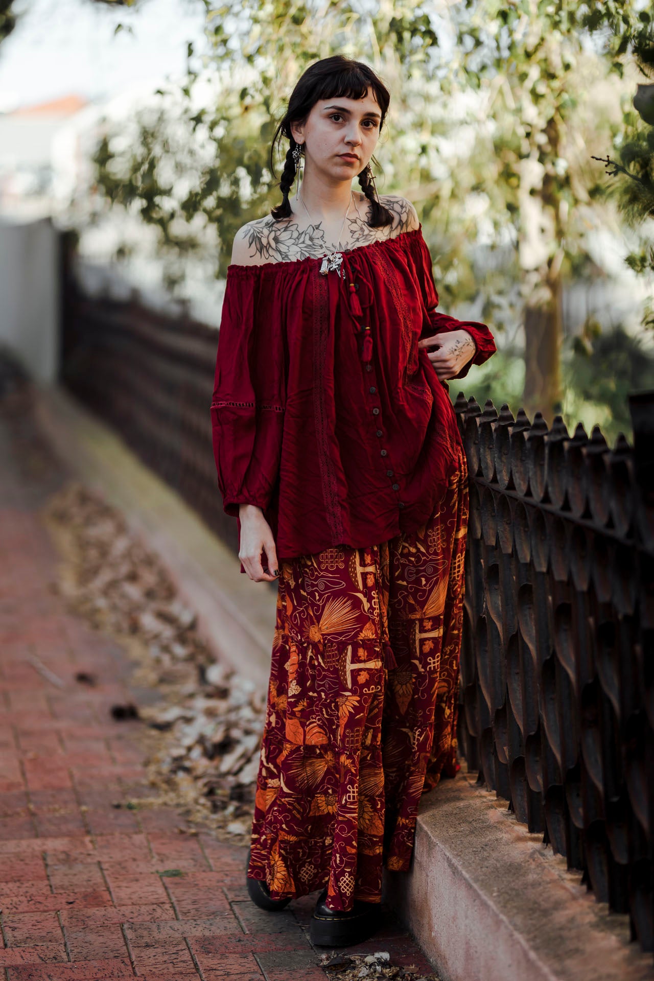 Woman in a red blouse and patterned pants standing against a fence with trees in the background