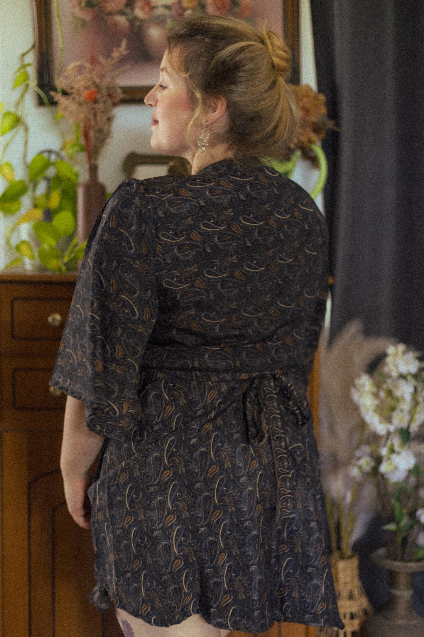 Woman wearing a dark patterned dress in an indoor setting with plants and furniture.