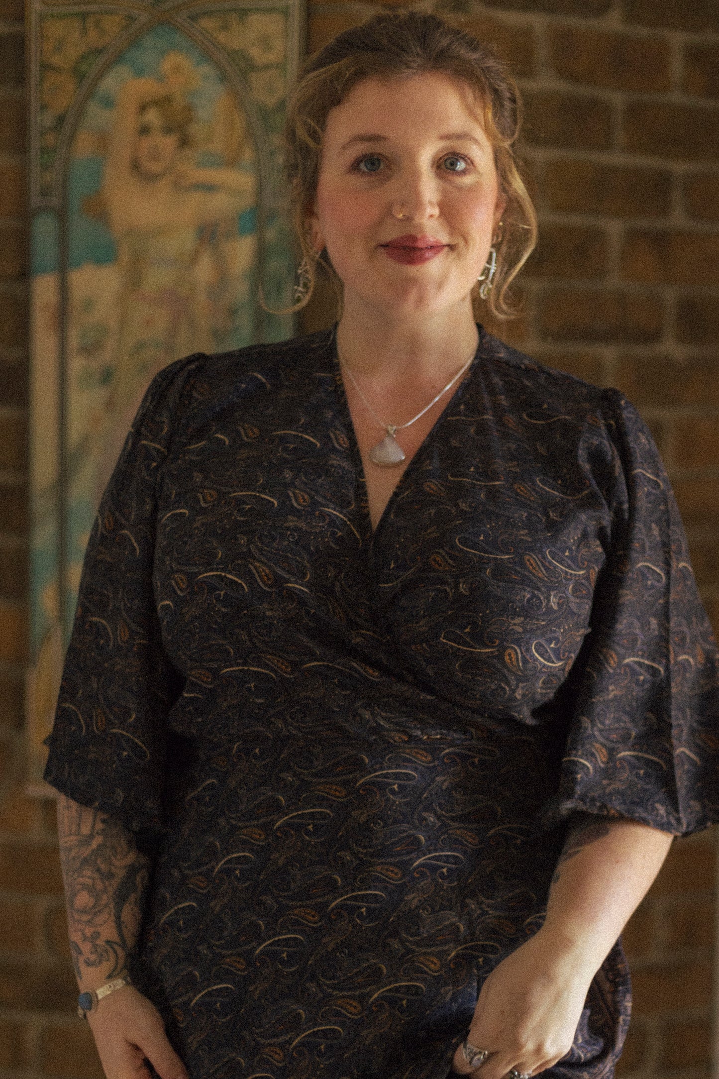 Woman wearing a dark patterned dress standing in front of a brick wall with decorative elements.