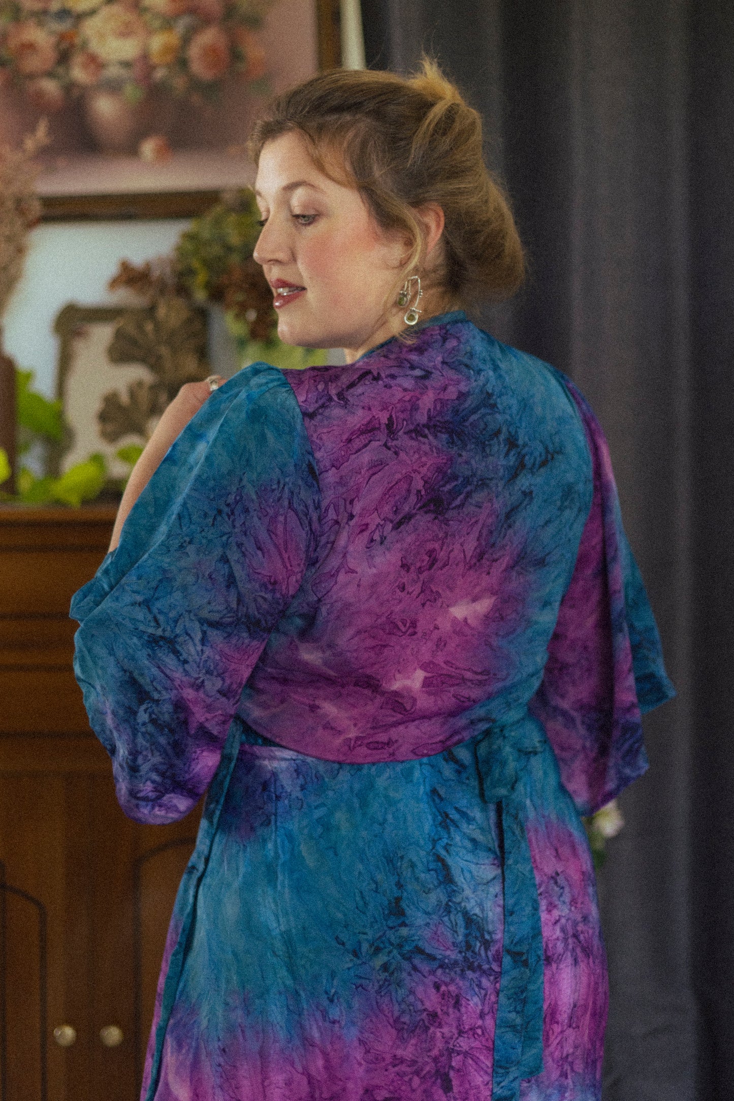 Woman wearing a blue and purple patterned dress in an indoor setting with a painting in the background.