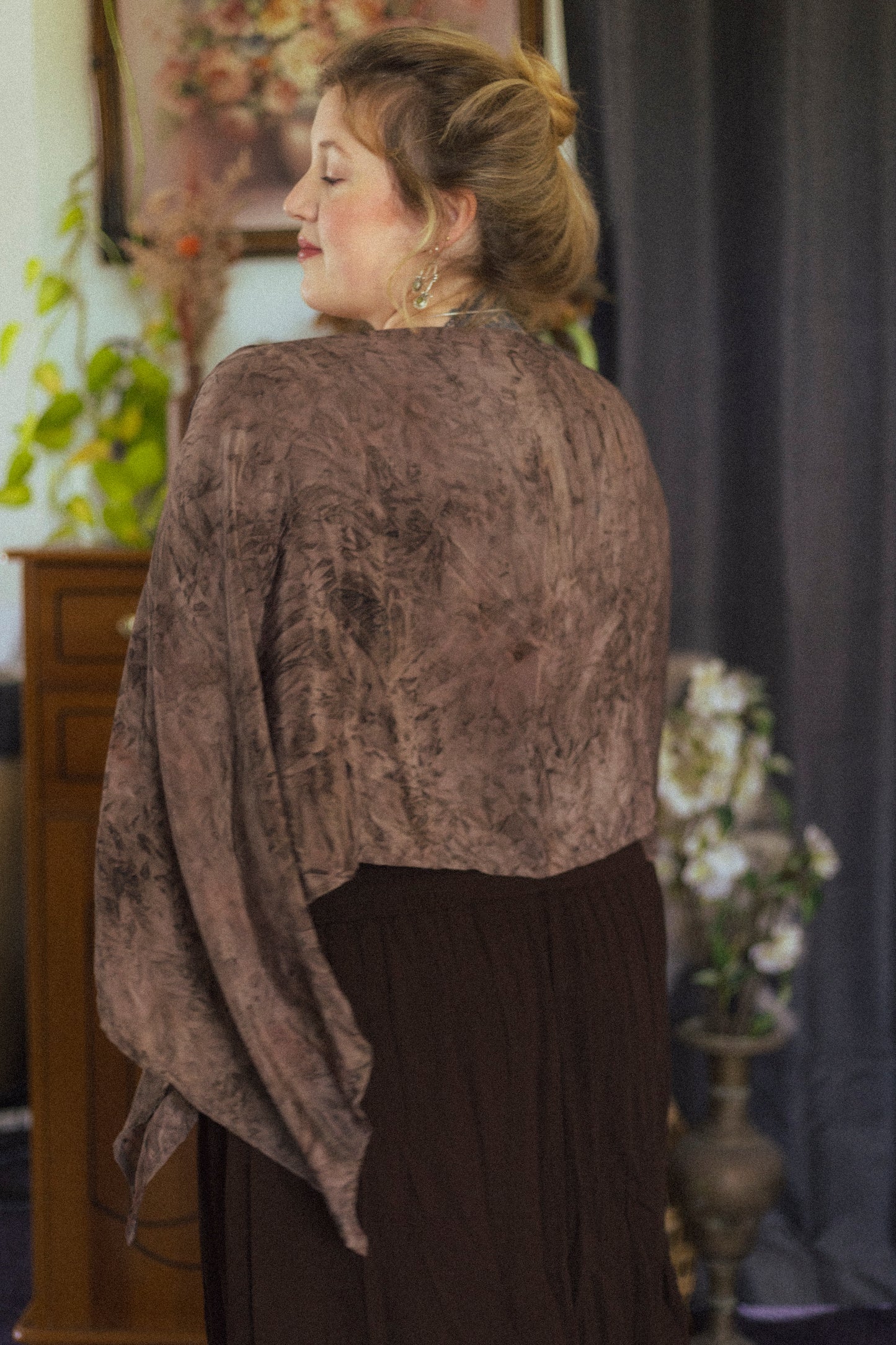 Woman wearing a brown textured shawl in an indoor setting with flowers and a mirror.