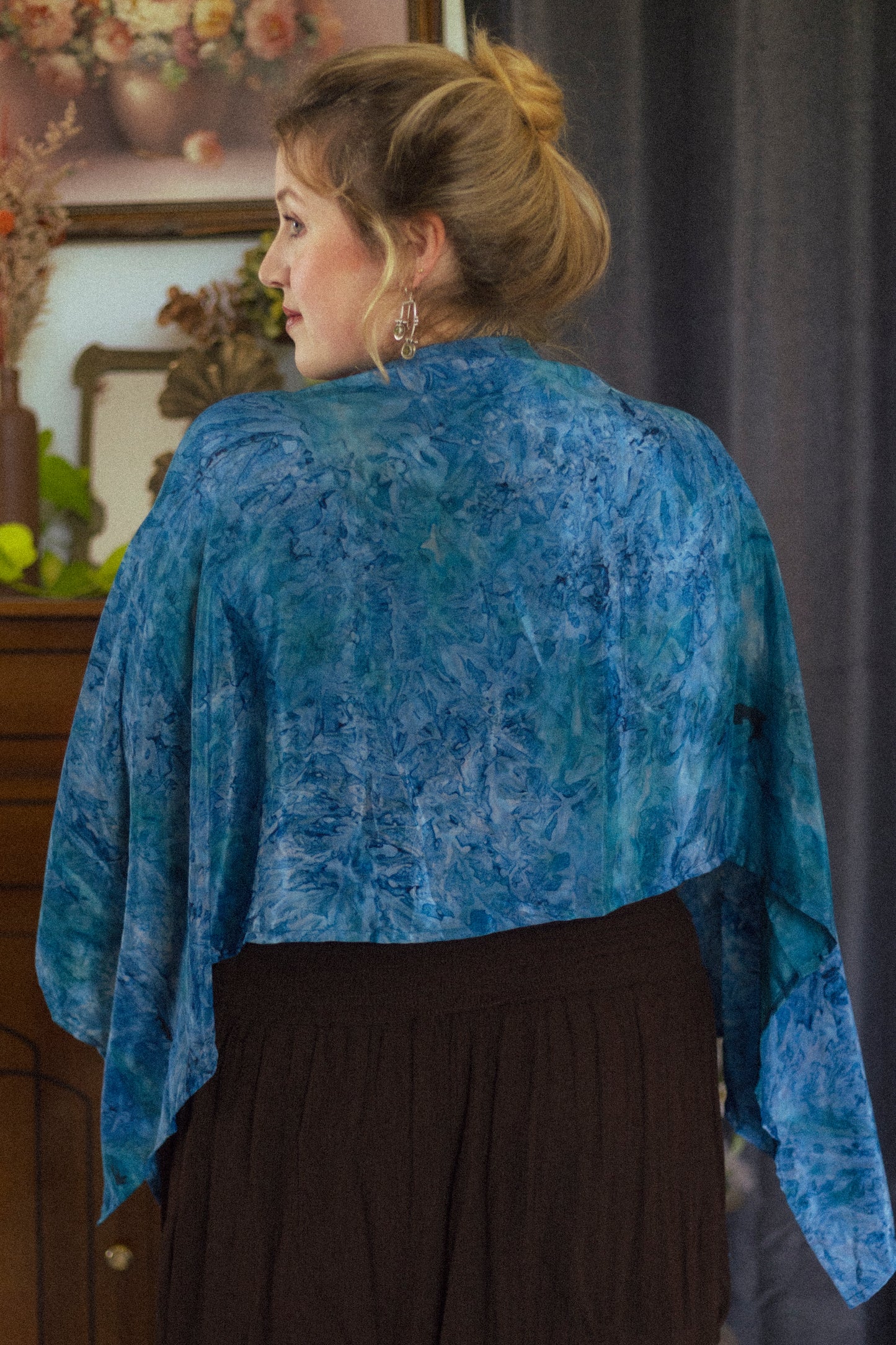 Woman wearing a blue patterned shawl over a dark dress, standing indoors with a blurred background.
