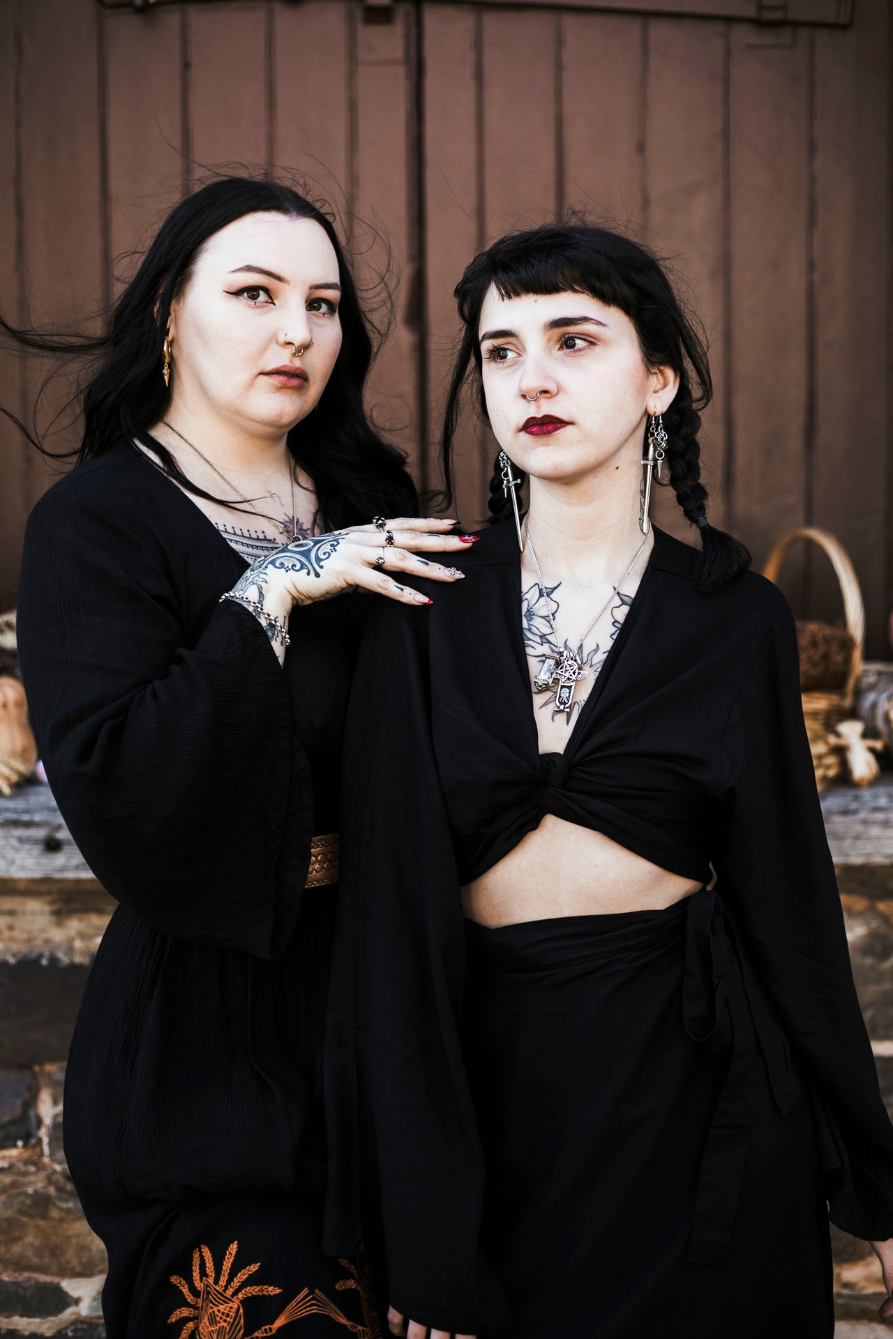 Two women in black attire with tattoos standing against a wooden wall.