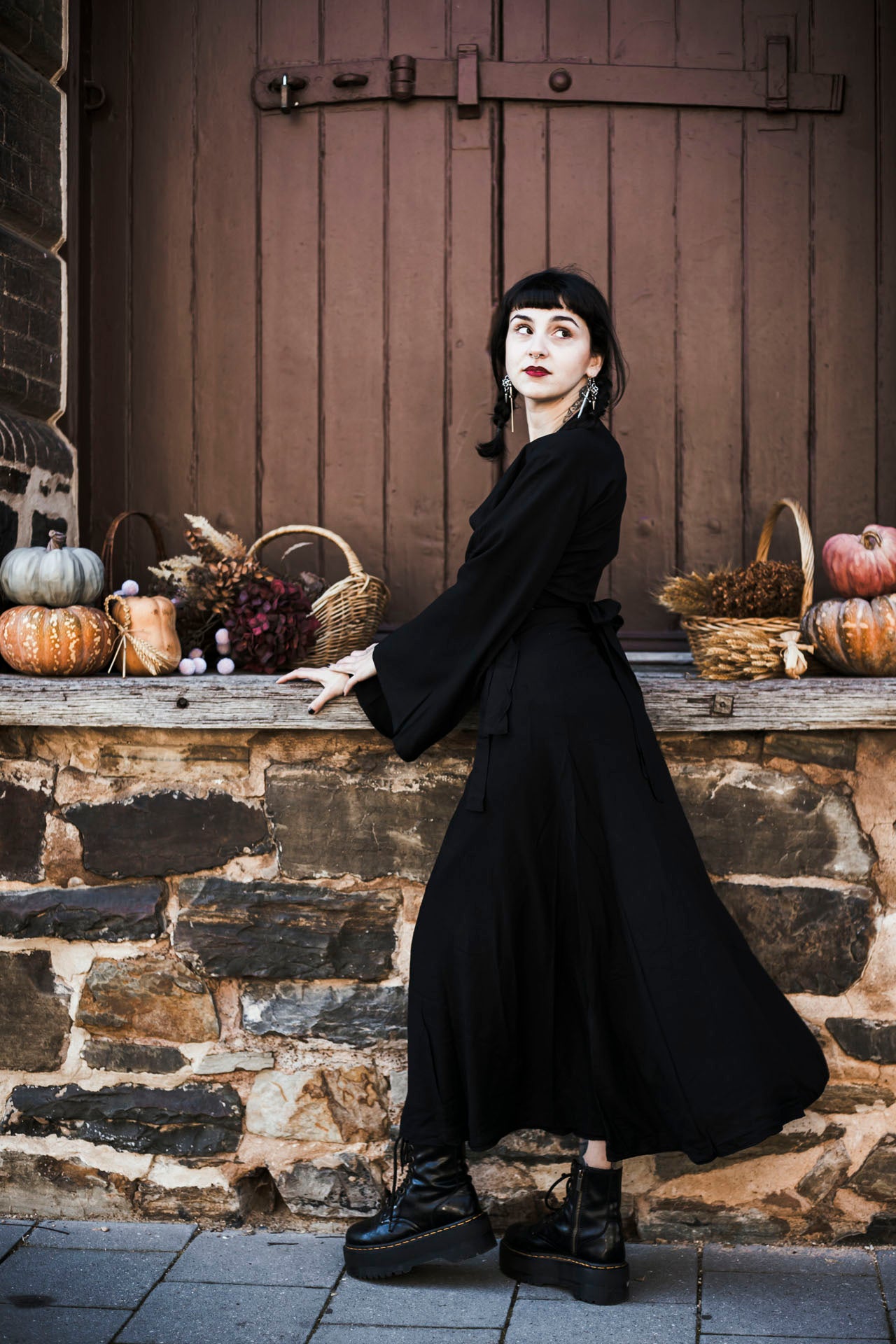 Woman in a long black coat standing in front of a stone wall with pumpkins and baskets.