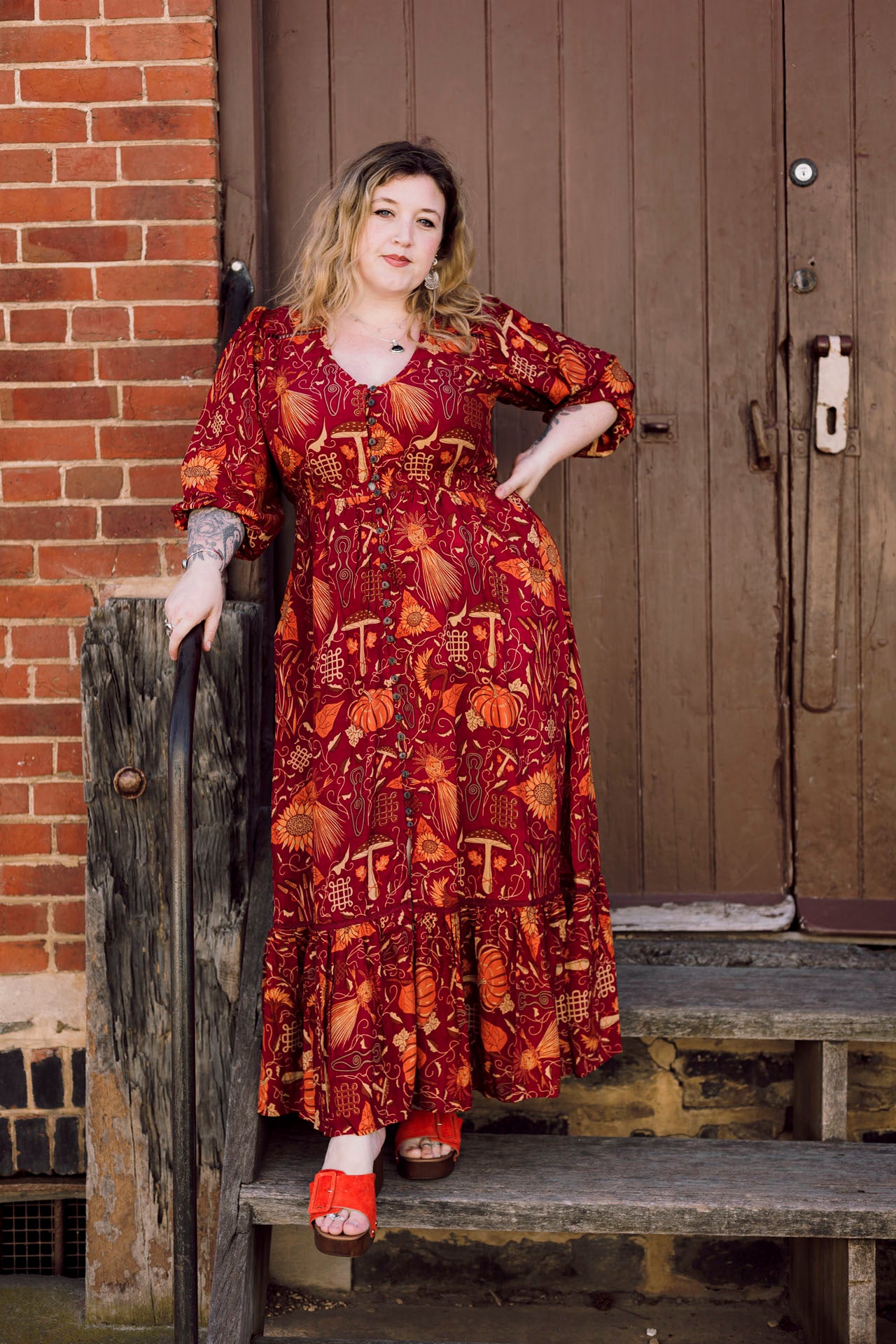 Woman in a red patterned dress standing on wooden steps against a brick wall.