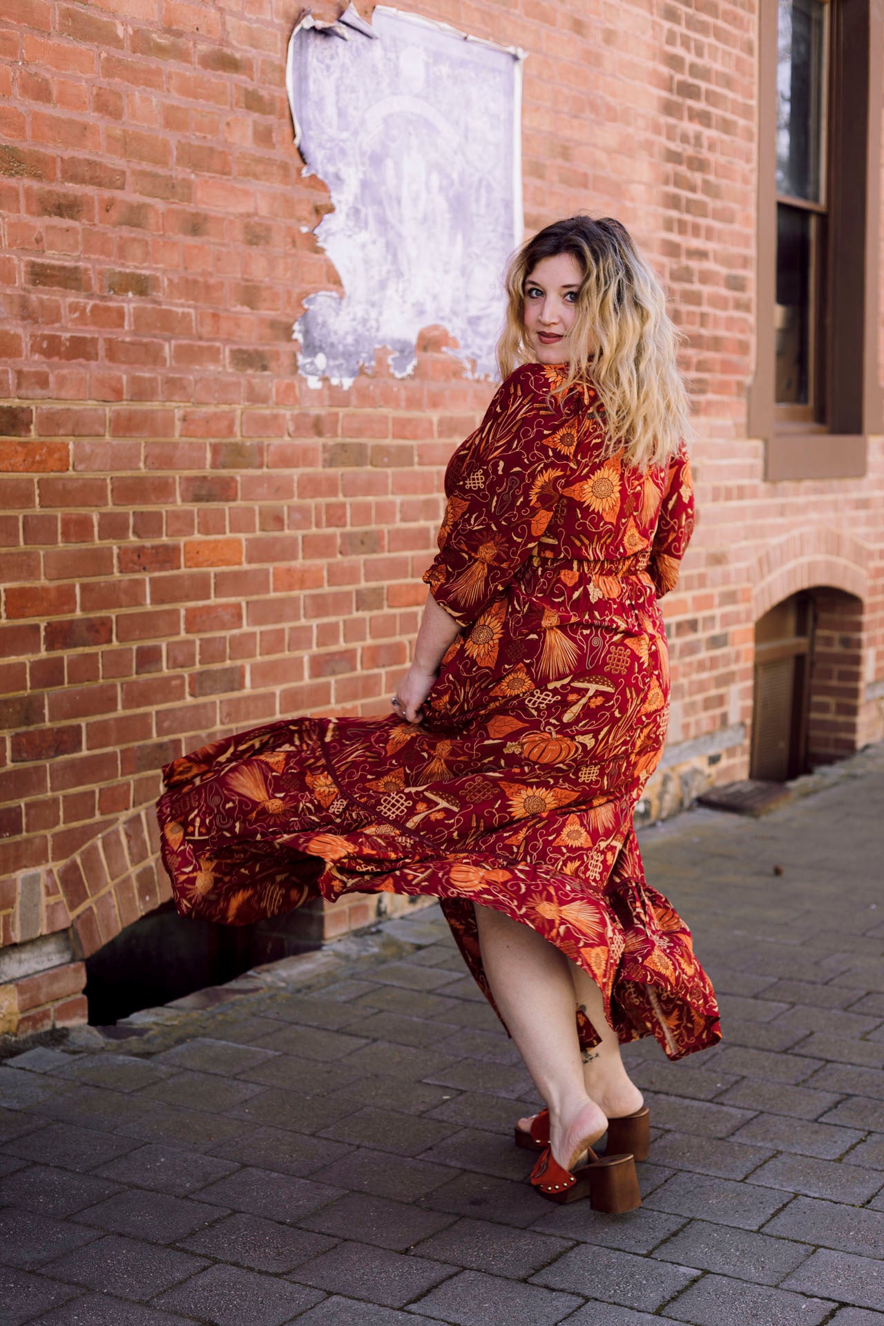 Woman in a red patterned dress posing against a brick wall.