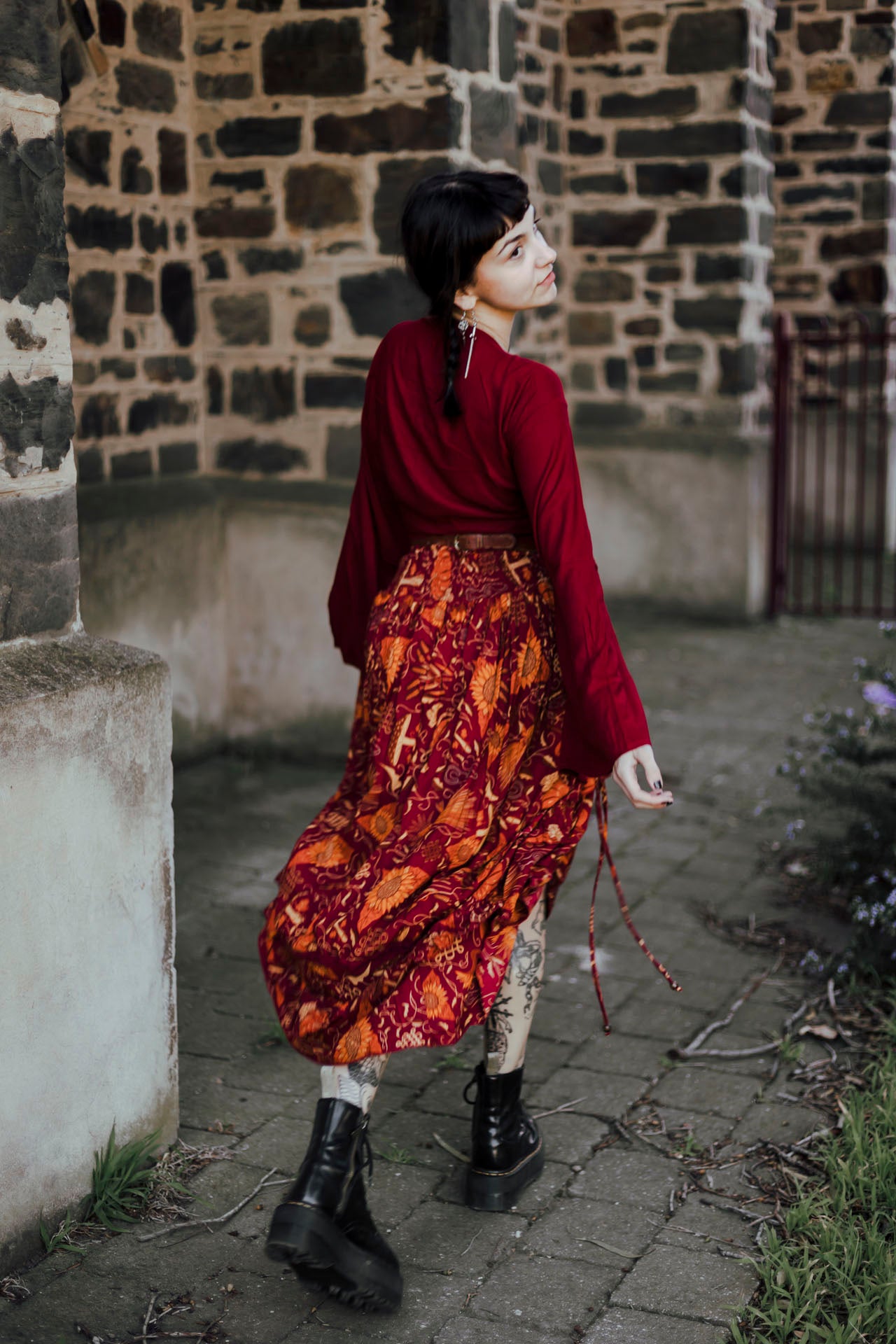 Person wearing a red top and patterned skirt standing against a stone wall.