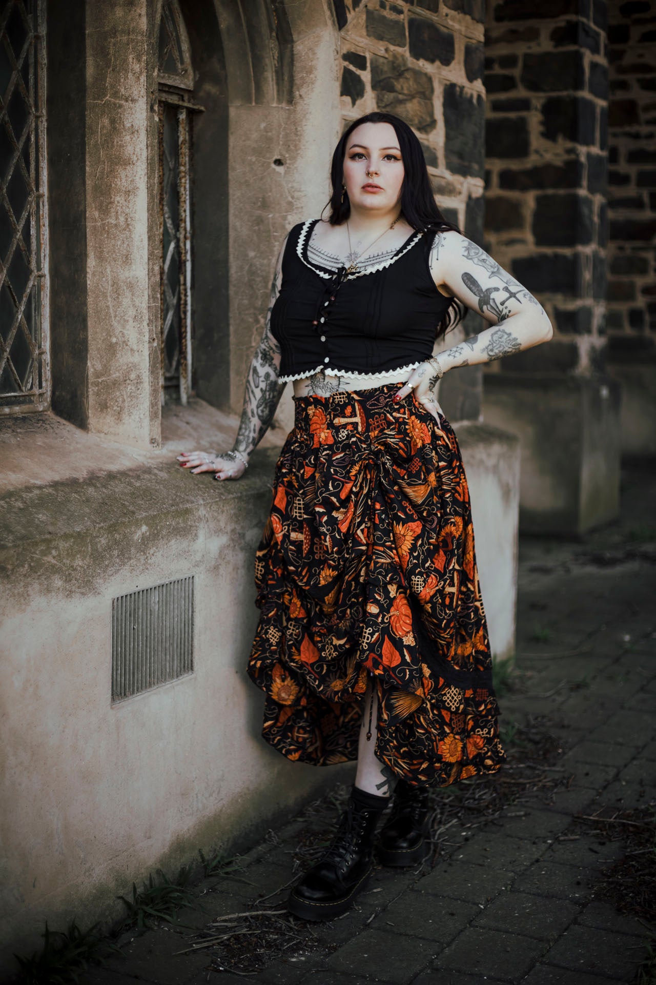 Person wearing a black top and patterned skirt standing against a stone wall.
