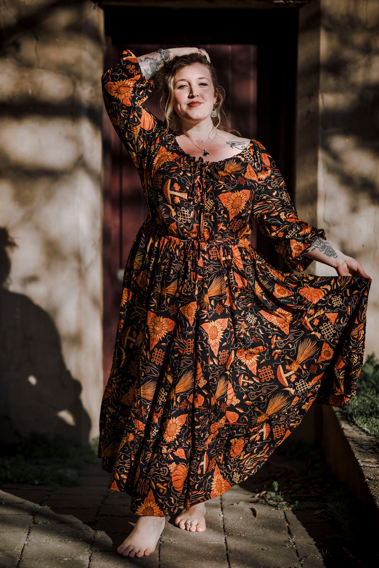 Woman in a long, patterned dress standing outdoors with a stone wall and wooden door in the background.