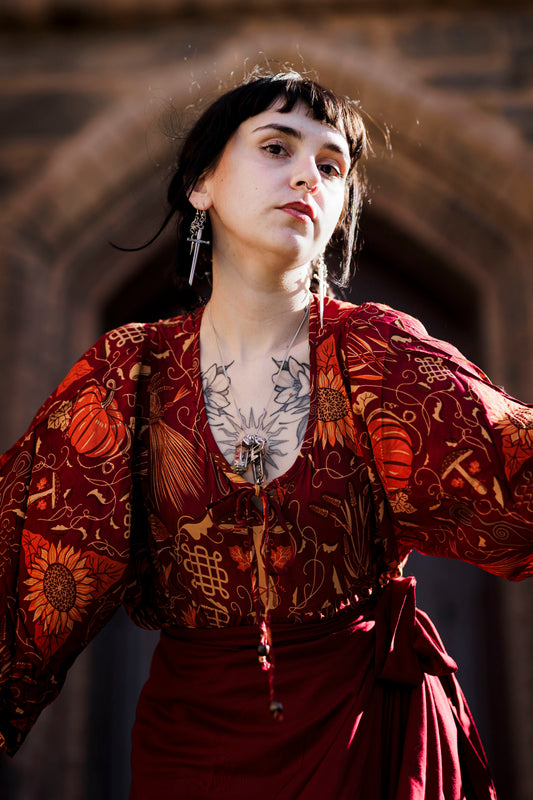 Woman in a patterned red garment with tattoos, standing against a stone archway.