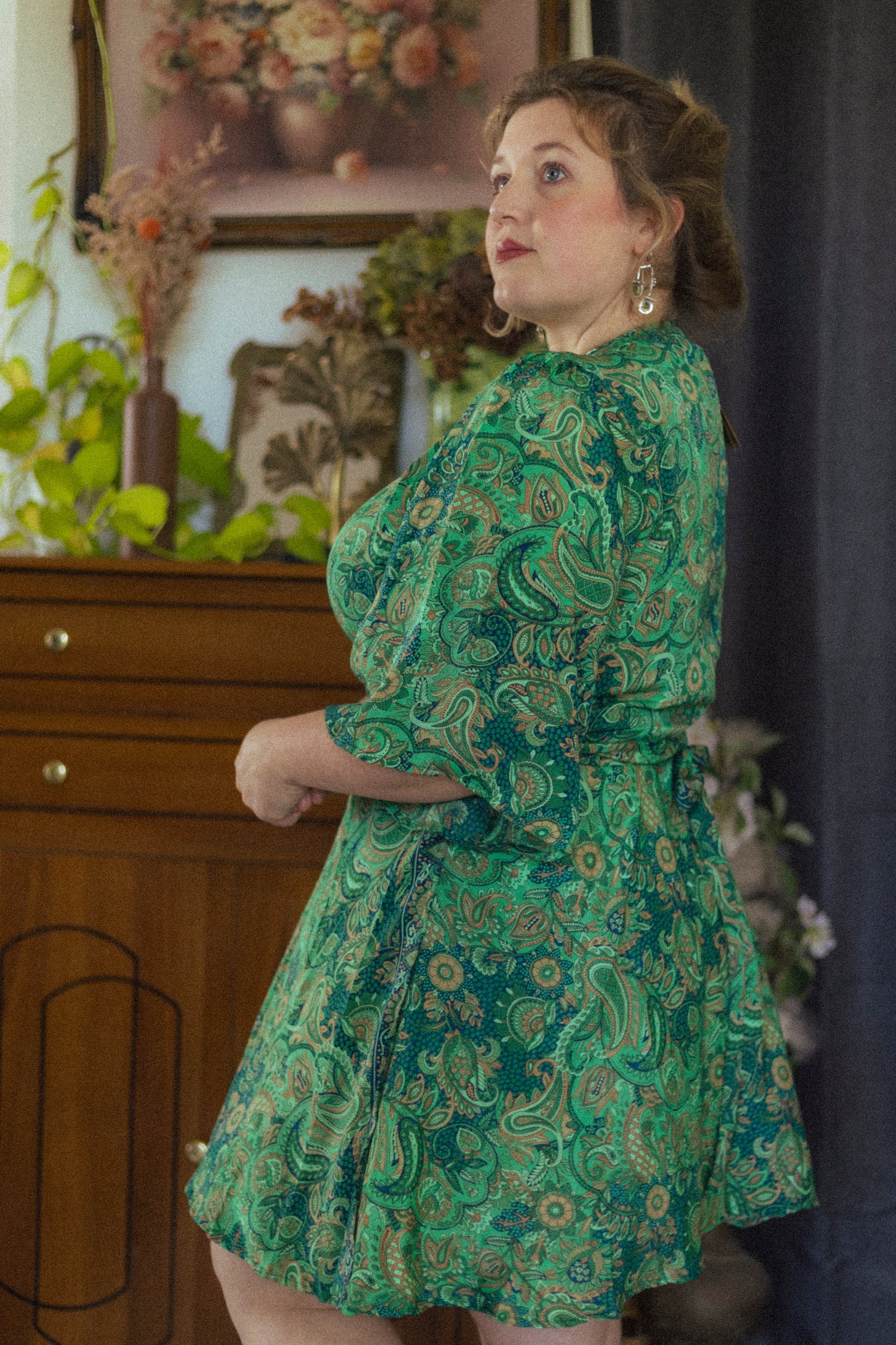 Woman wearing a green paisley dress standing in a room with decorative elements.
