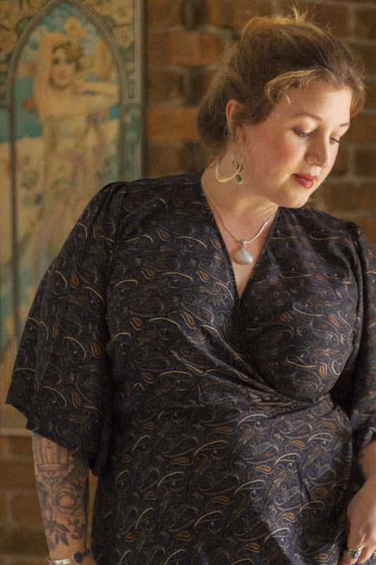 Woman wearing a dark patterned dress in front of a brick wall with decorative elements.