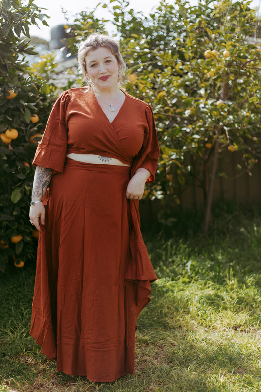 Woman in a long rust-colored dress standing in an outdoor setting with greenery and fruit trees.