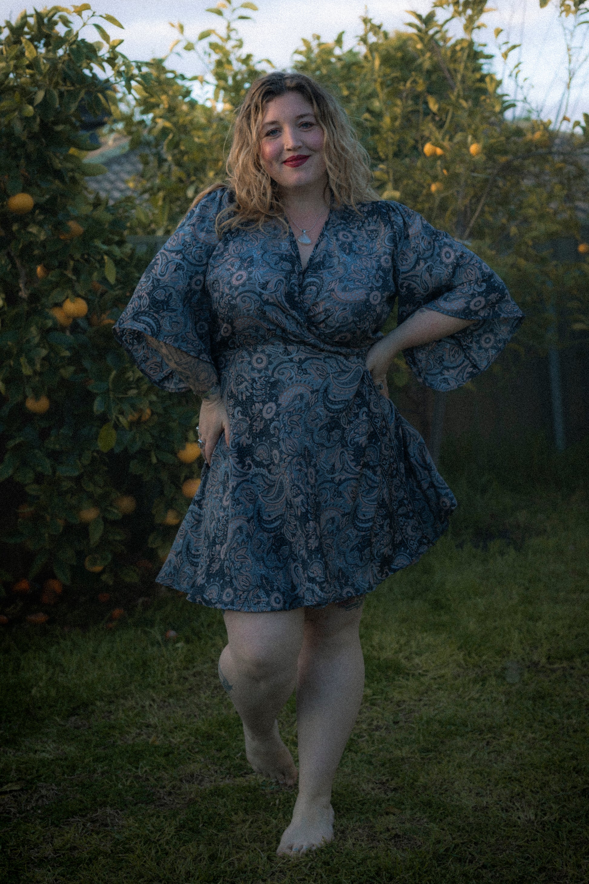 Woman in a blue floral dress standing in an outdoor setting with greenery and fruit trees.