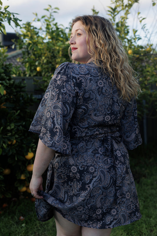 Woman wearing a dark blue patterned dress standing outdoors with greenery in the background