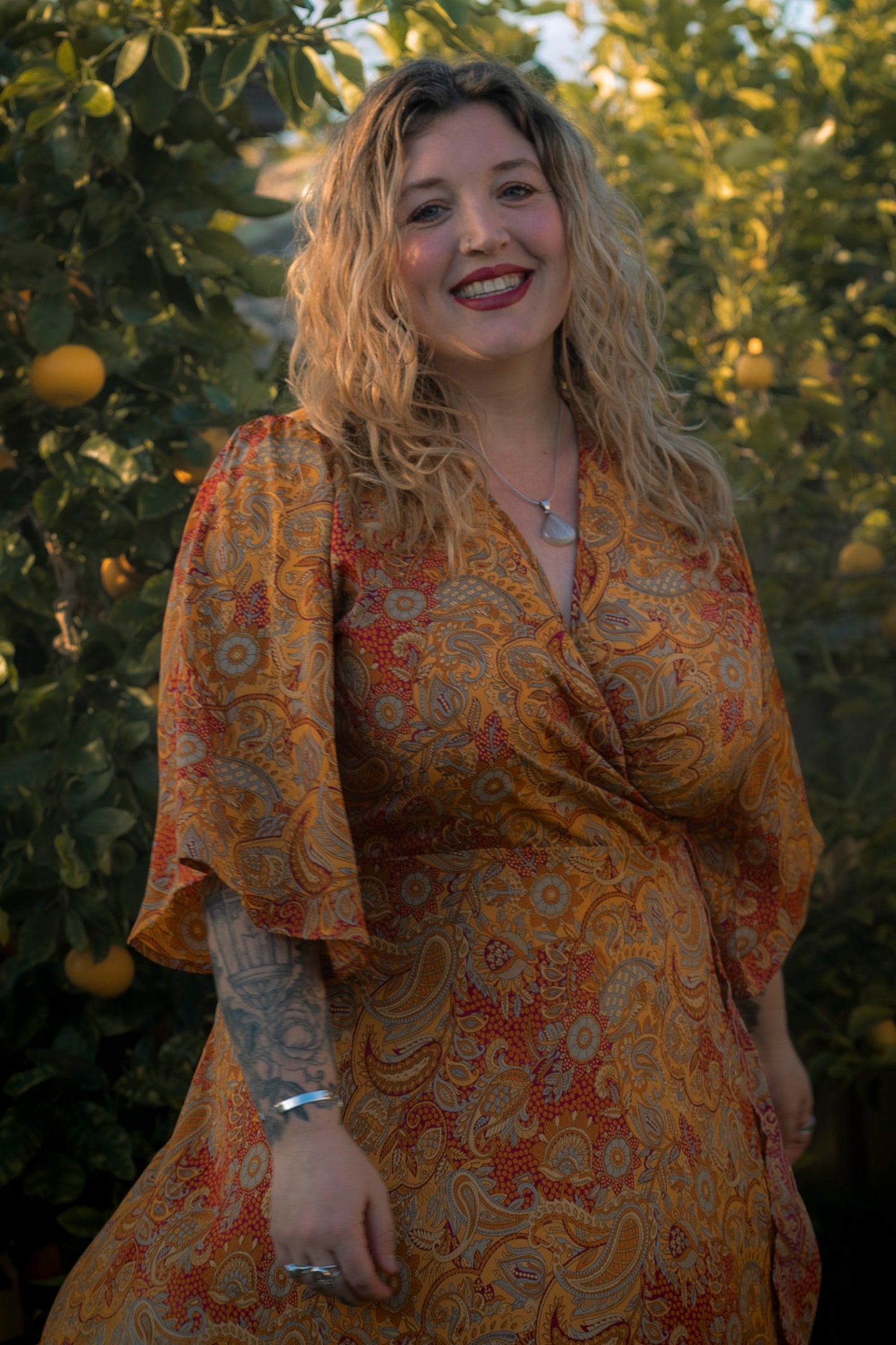 Woman wearing a patterned orange dress standing in front of green foliage.