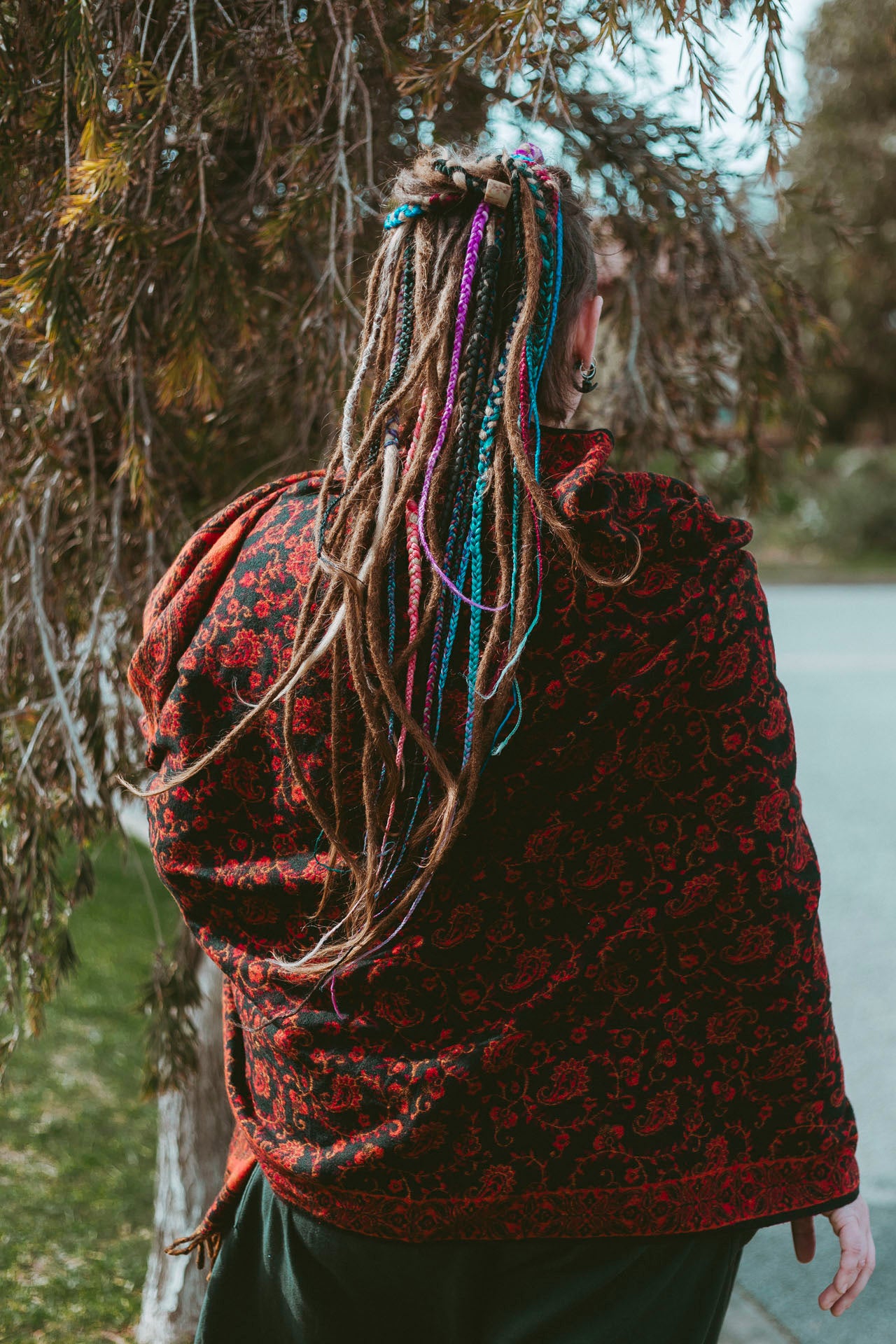 Person wearing a patterned red and black garment with colorful braids, standing outdoors near trees.