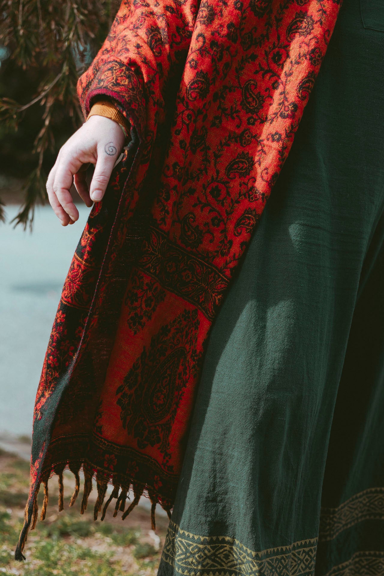 Person wearing a red patterned shawl standing outdoors near trees and water.